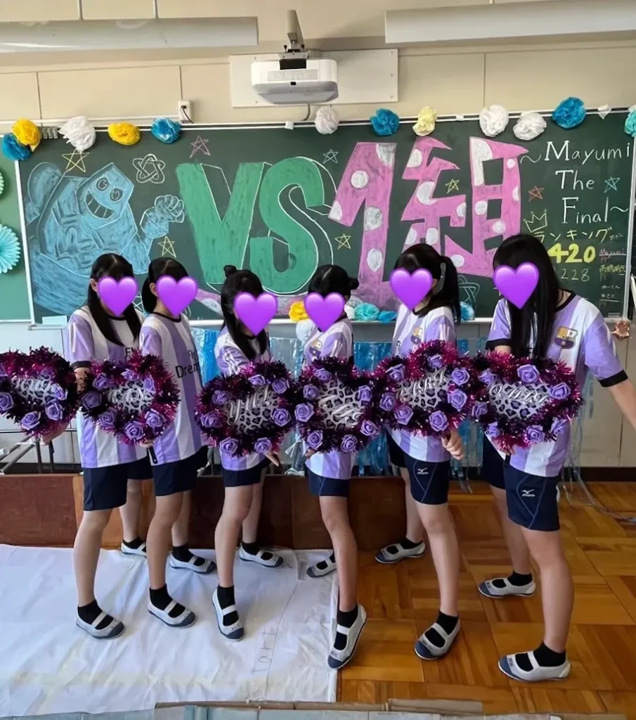 紫色のサッカーユニフォーム風クラスTシャツを着ながら、ヒョウ柄と紫色のバラと黒と紫のキラキラモールでデコったうちわを手に持って黒板の前で写真を撮っている女子高生6人